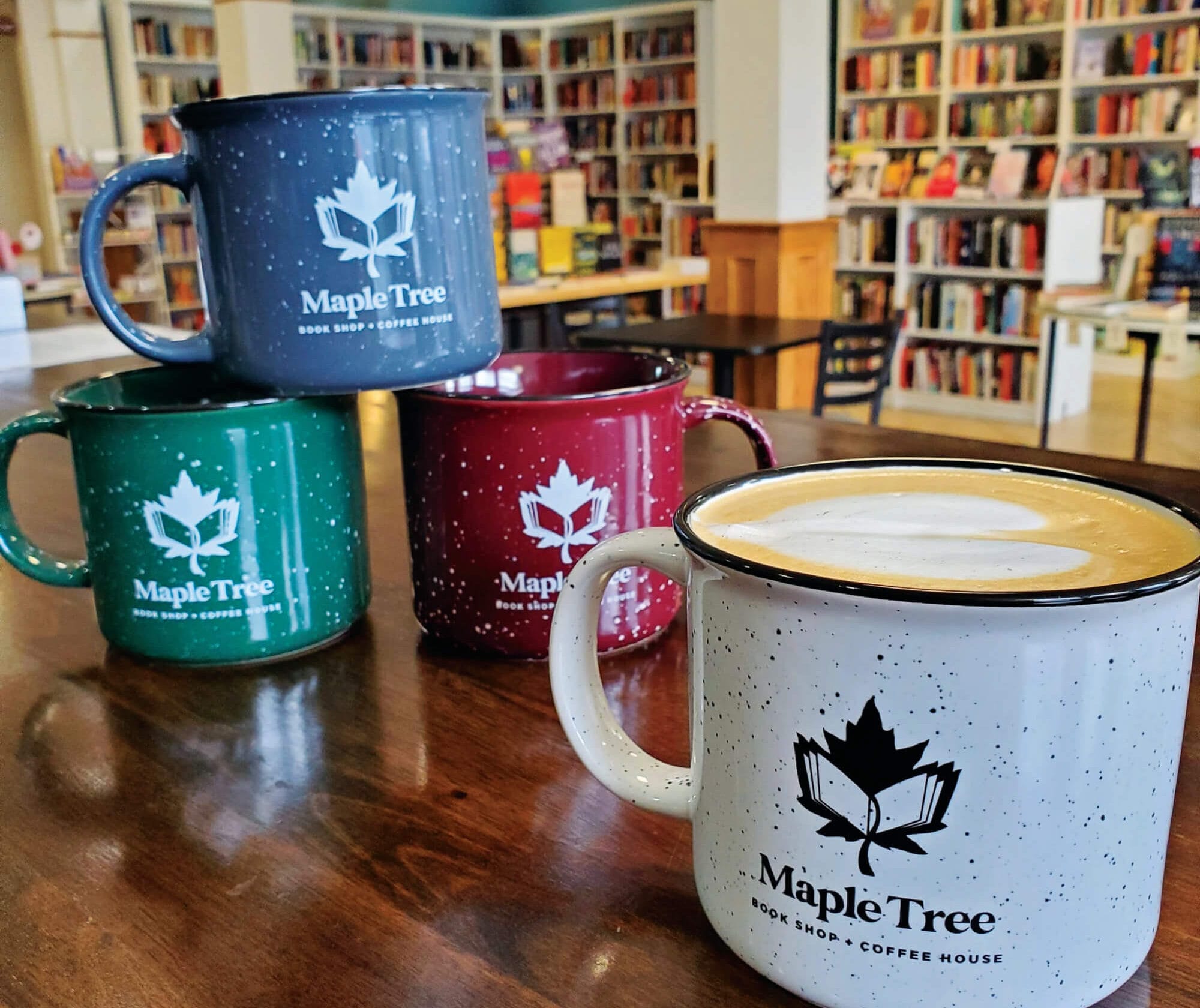 A Maple Tree Book Shop + Coffee House mug filled with hot coffee on a table with bookshelves lining the back wall.